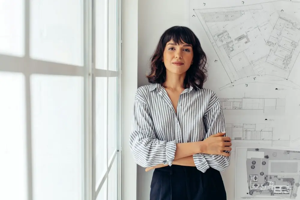 Woman standing by window in office with building blueprints on the wall behind her