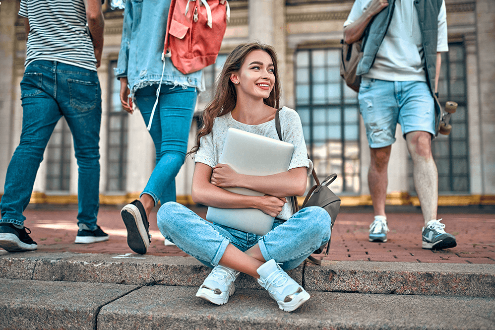 A charming girl student with a backpack and a laptop sits on the steps near the campus against the background of students passing by, reflecting student banking at Northern Birch