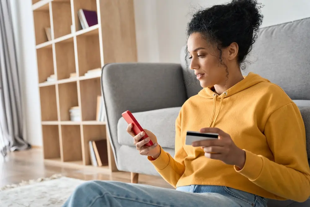 A woman relaxing on the living room floor holding a credit card and her phone