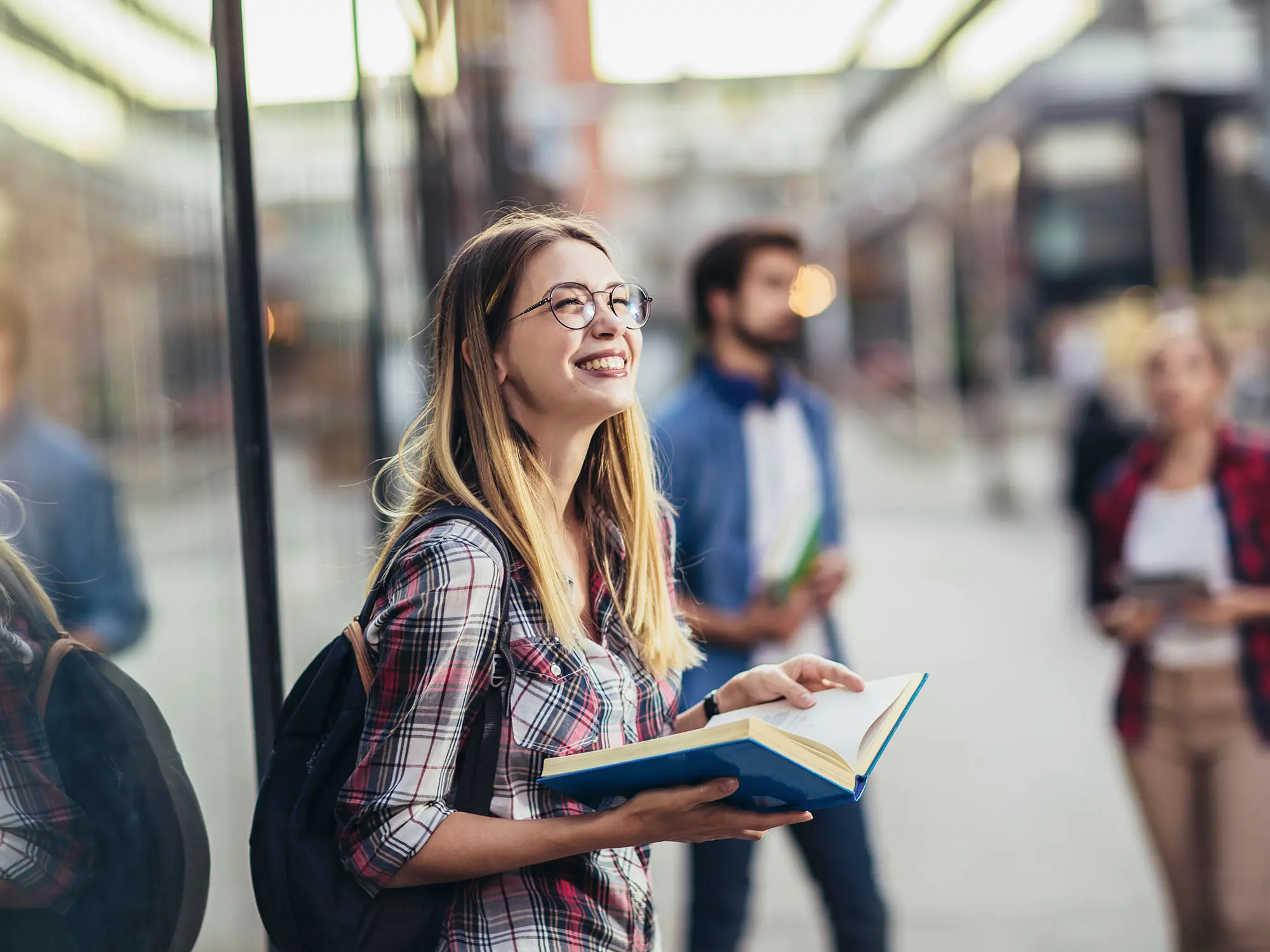 A young woman holding a textbook and wearing a backpack smiling